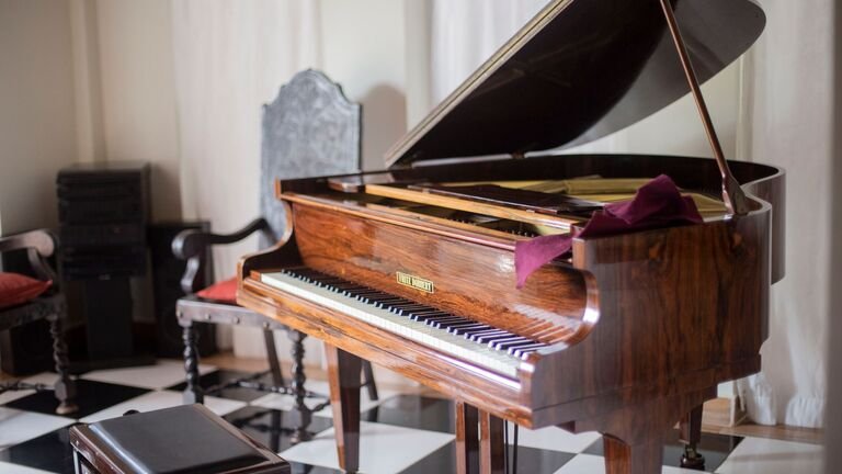 Piano de cola de madera con su banqueta en un salón de suelo ajedrezado, antes de prepararlo para el traslado